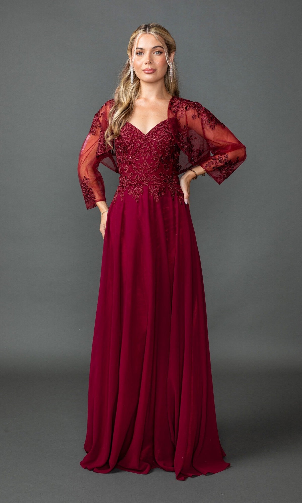 A woman models the Dancing Queen 4542 long prom dress, featuring sheer lace embroidered sleeves, against a plain gray background.